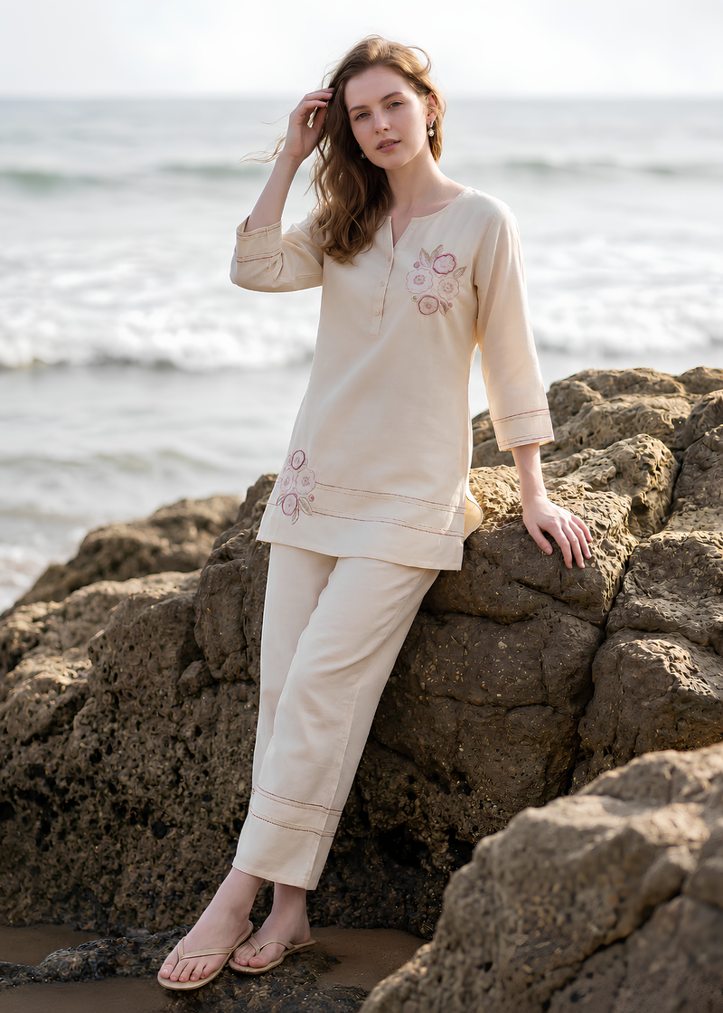 A woman wearing a cream-colored two-piece kurta set with pink floral embroidery, posing on large brown rocks with the ocean in the background.